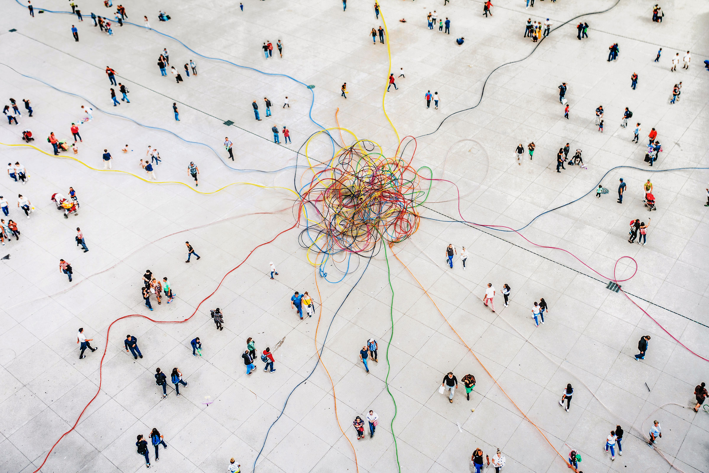 cirQum-beratung-datenintegritaet Blick aus der Vogelperspektive auf ein riesiges buntes Kabelknäuel, von dem aus einzelne Kabel in alle Richtungen laufen, auf einem Platz voller Menschen.