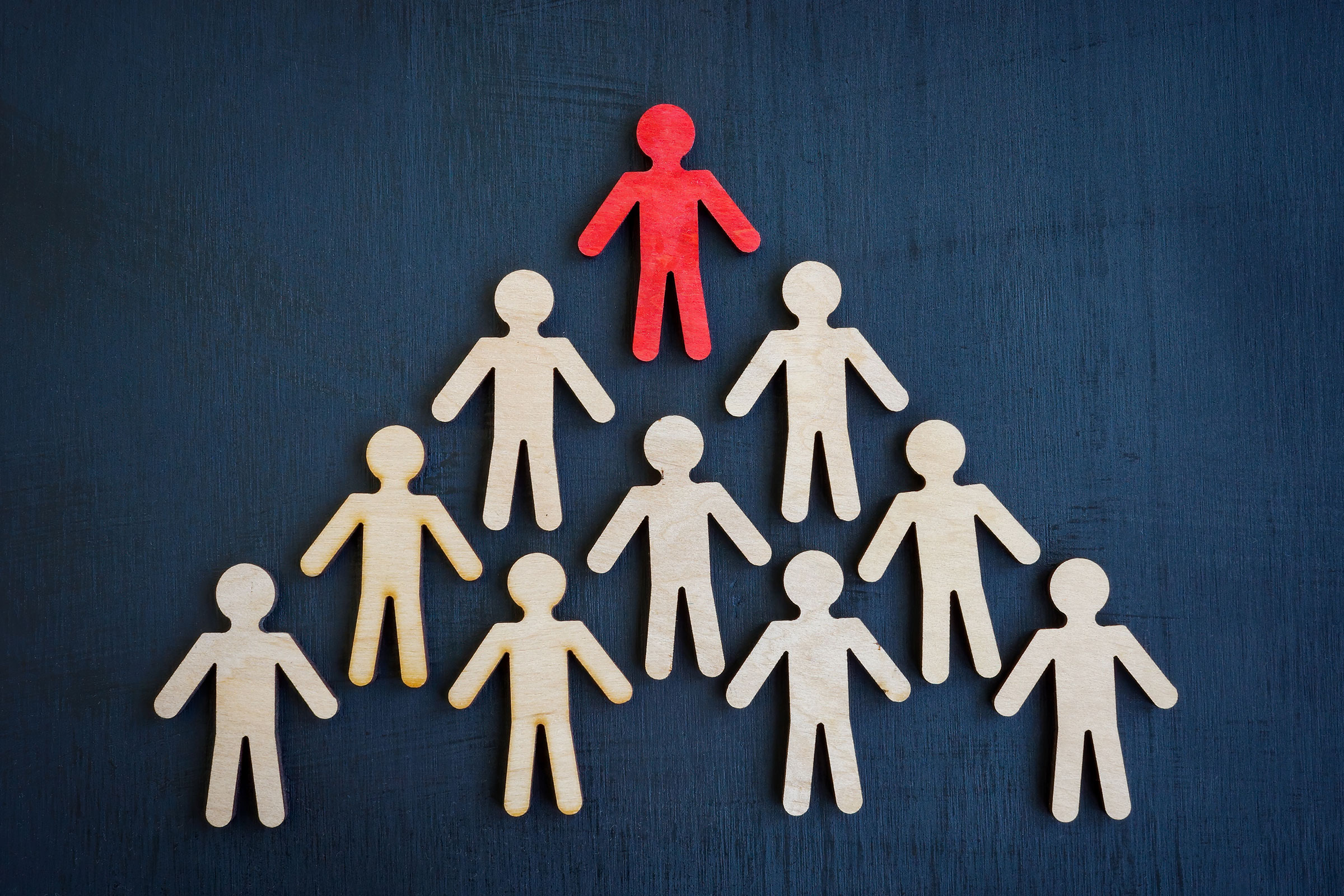 cirQum-schulungen-der-kleine-grosshandel Zehn aus Holz ausgeschnittene menschliche Figuren in einer Pyramide angeordnet. Die oberste Figur ist rot, die anderen naturfarben.