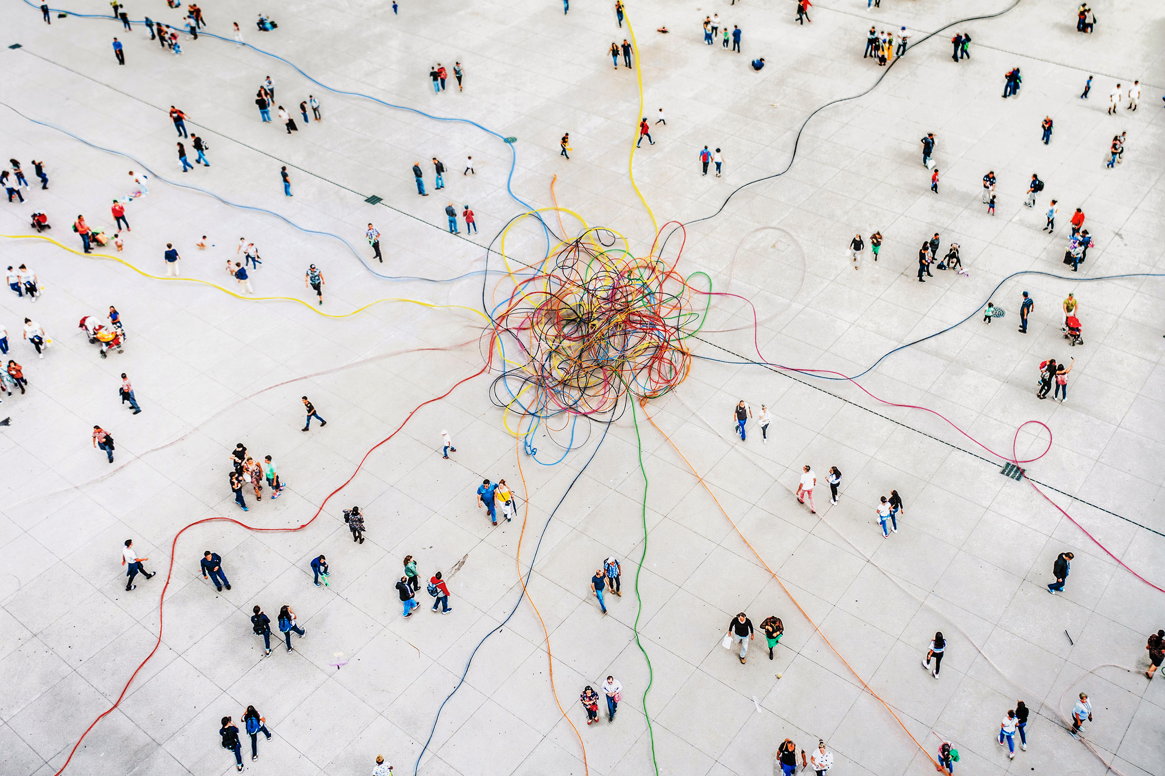 cirQum-schulungen-dokumentationdatenintegritaet Blick aus der Vogelperspektive auf ein riesiges buntes Kabelknäuel, von dem aus einzelne Kabel in alle Richtungen laufen, auf einem Platz voller Menschen.